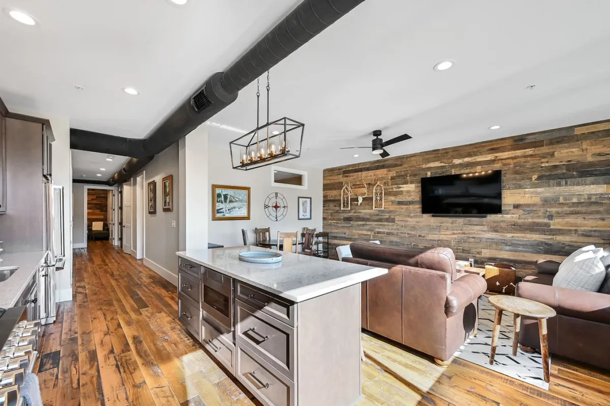 This image reveals a spacious, open-concept kitchen and living area, featuring a central kitchen island set against a striking wooden accent wall. The seating area is stylishly appointed, anchored by a contemporary chandelier that adds a touch of boutique elegance. The cozy atmosphere invites guests to relax and socialize after a day out.