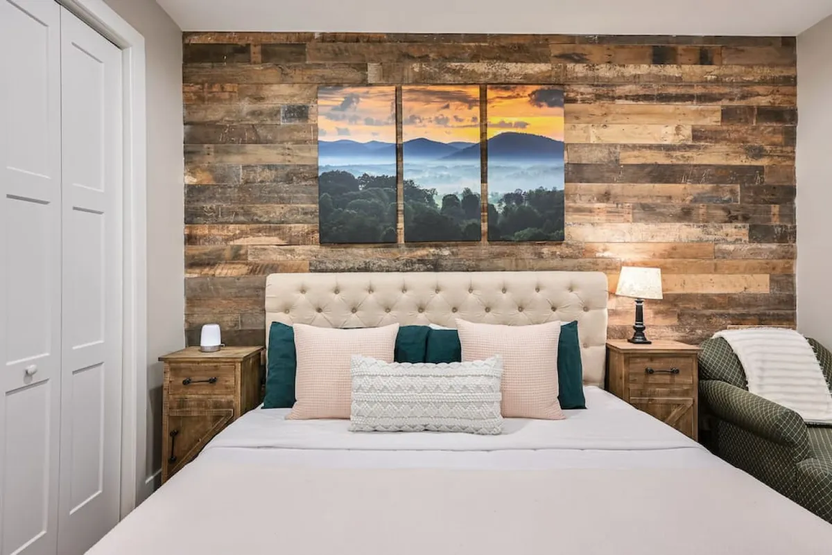 A serene king bedroom featuring a tufted headboard, landscape artwork, a nightstand with a lamp, and decorative pillows. The textured wall and soft color palette create an inviting, boutique-style atmosphere ideal for relaxation and unwinding.