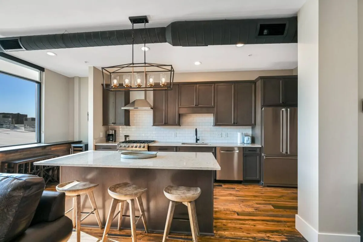 This image features a spacious kitchen with a prominent island countertop, wooden stools, modern appliances, and large windows that allow natural light to pour in. The clean lines and contemporary fixtures highlight the kitchen’s upscale feel, while the seating at the island invites socializing and casual meals.
