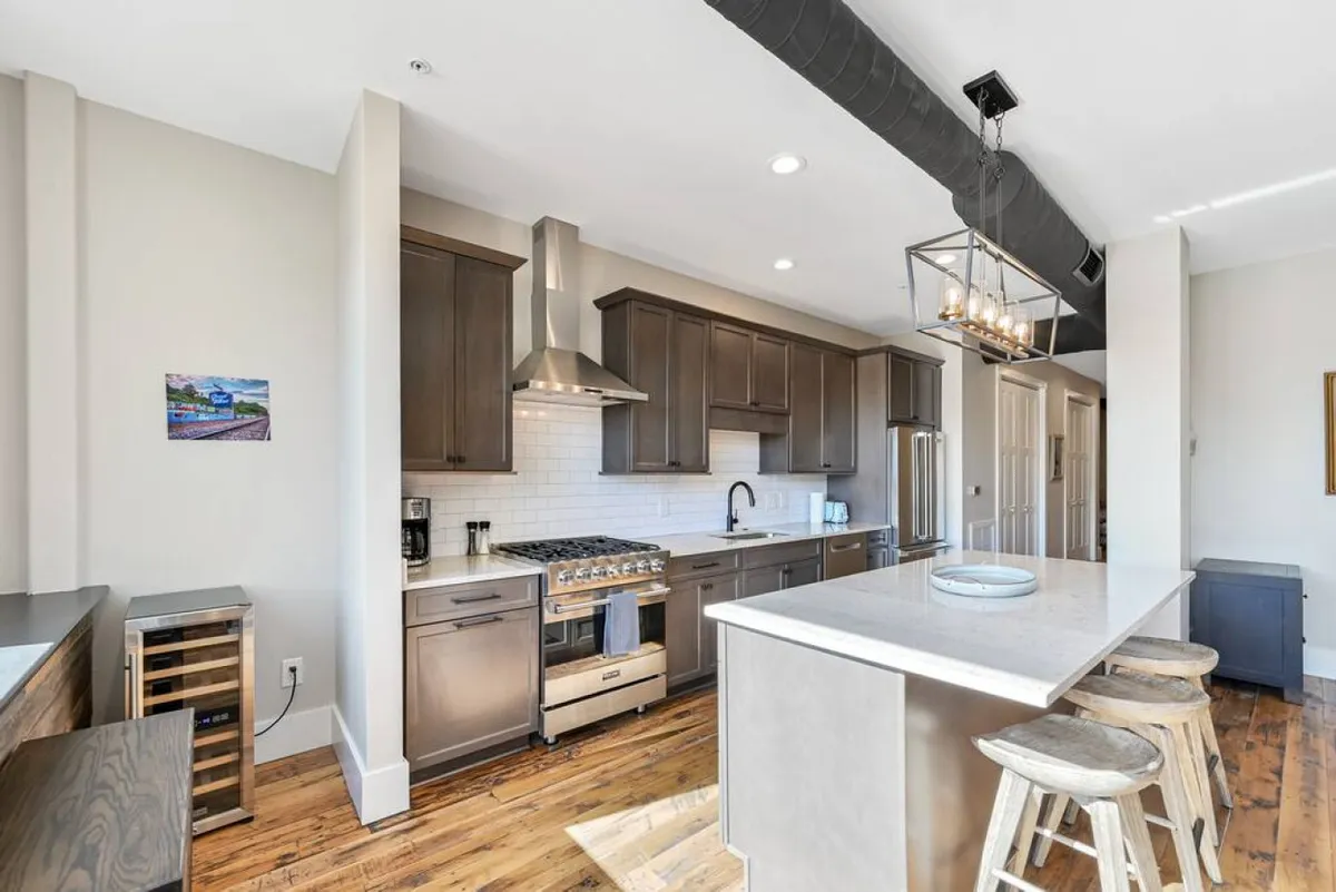This image shows a kitchen with a sleek island, stainless steel appliances, wooden cabinetry, and bar stools neatly arranged for casual dining or morning coffee. The overall design is contemporary yet warm, blending modern function with inviting comfort.
