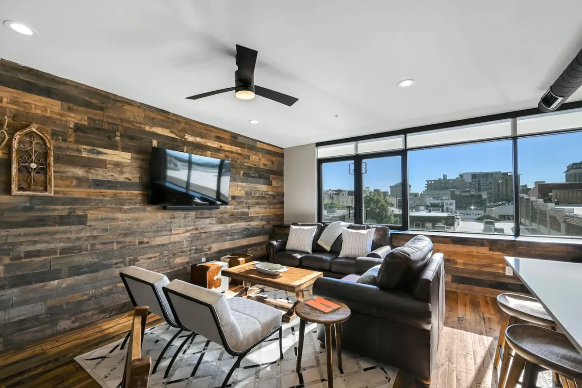 A cozy, inviting living room with comfortable seating, a modern TV, and large windows that offer a view outside. Decorative accents and a warm color palette add to the sense of comfort, while the windows subtly connect the space to the city beyond.