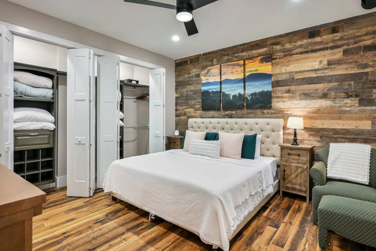 A plush, tufted king bed set against a wooden accent wall offers a glimpse into the serene bedroom space. A tasteful landscape artwork, soft nightstand lighting, and carefully curated decor evoke sophistication and intimacy, perfect for couples seeking a romantic getaway.