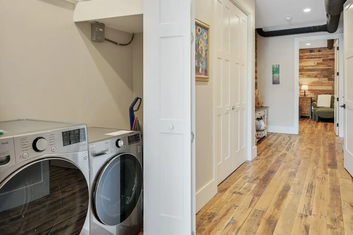 This image centers on a practical laundry area with a washer and dryer along a warm wooden floor, leading through a doorway to the next room. Artwork on the wall and the visible connection to the adjoining living space create a sense of functional flow within the property.