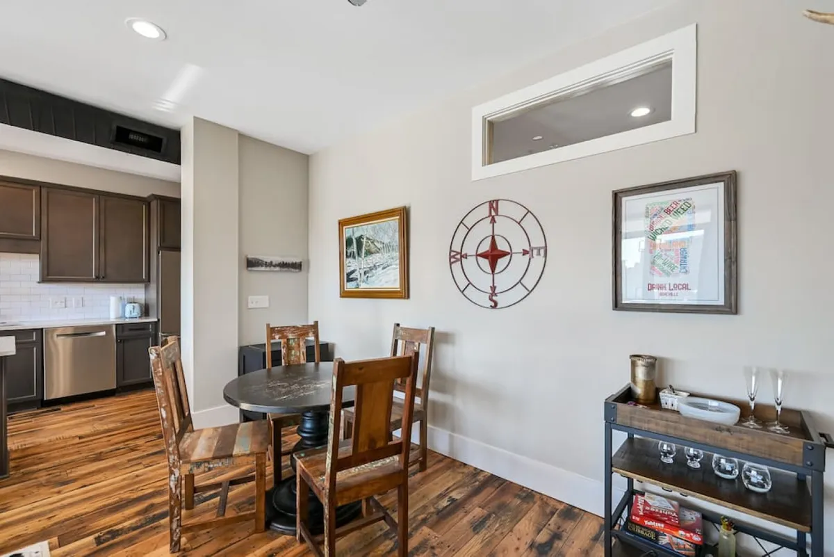 A well-set wooden dining table surrounded by matching chairs, with modern kitchen appliances visible in the background. Decorative wall art adds personality, and the scene is set for both casual meals and more formal gatherings.