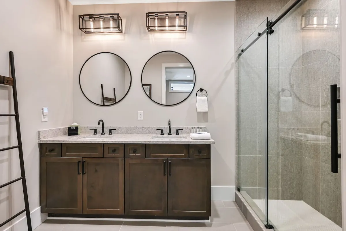 A spa-inspired ensuite bathroom with a double vanity featuring dark cabinetry, two circular mirrors, and a glass-enclosed shower. The space feels serene and sophisticated, perfect for morning routines or evening relaxation.