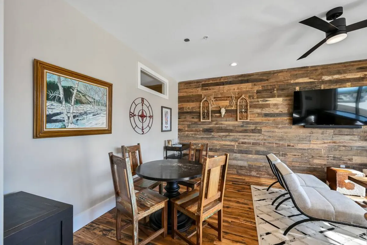 A cozy dining and living area featuring wooden wall paneling, an inviting dining table, curated artwork, a television, and a ceiling fan. The scene exudes warmth and a sense of relaxation, emphasizing spaces for socializing or recharging after a day out.