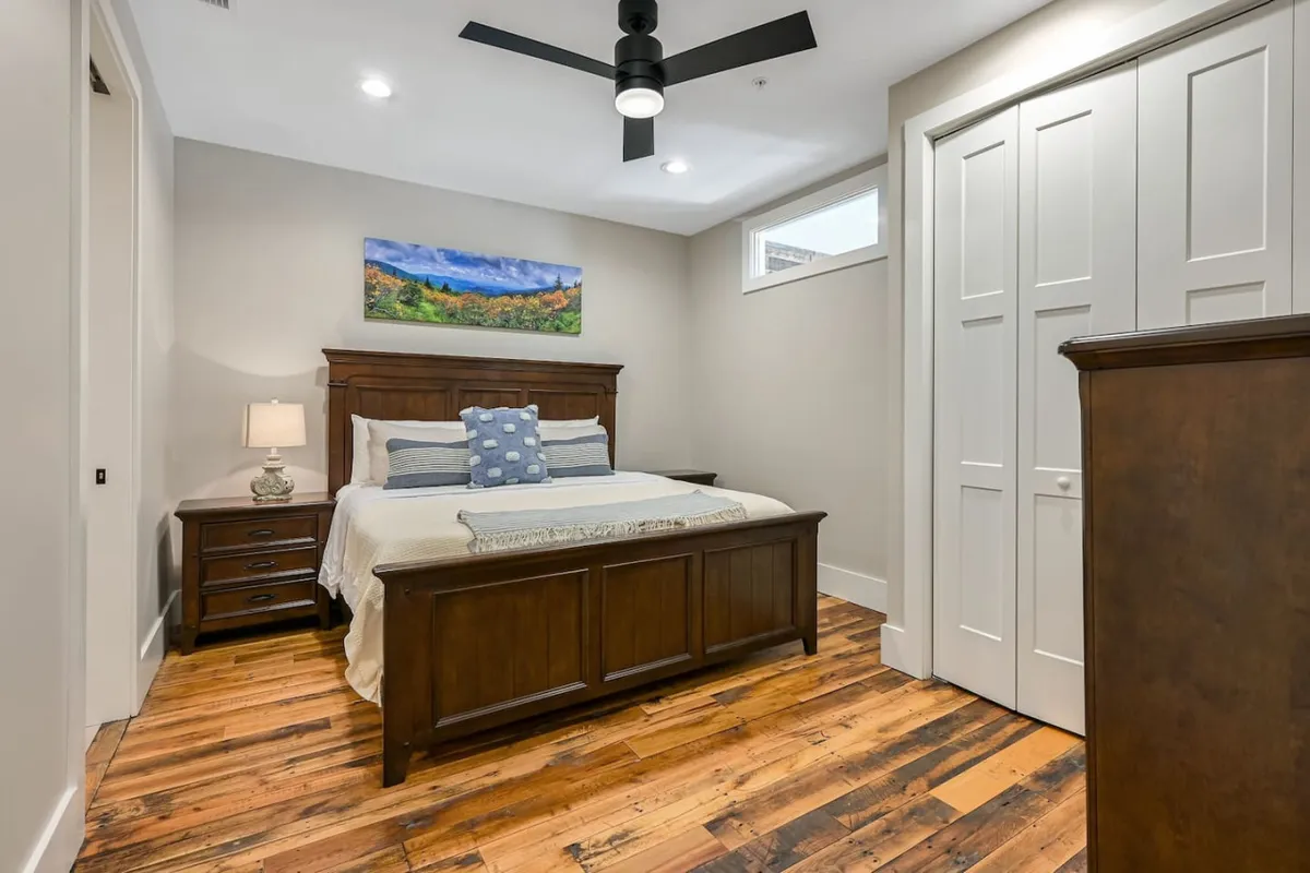 This image features a king-size wooden bed frame adorned with blue decorative pillows, a nightstand with a lamp, and a calming landscape painting above the bed. The overall mood is cozy and peaceful, with natural light streaming in, highlighting the plush bedding and comfortable ambiance.