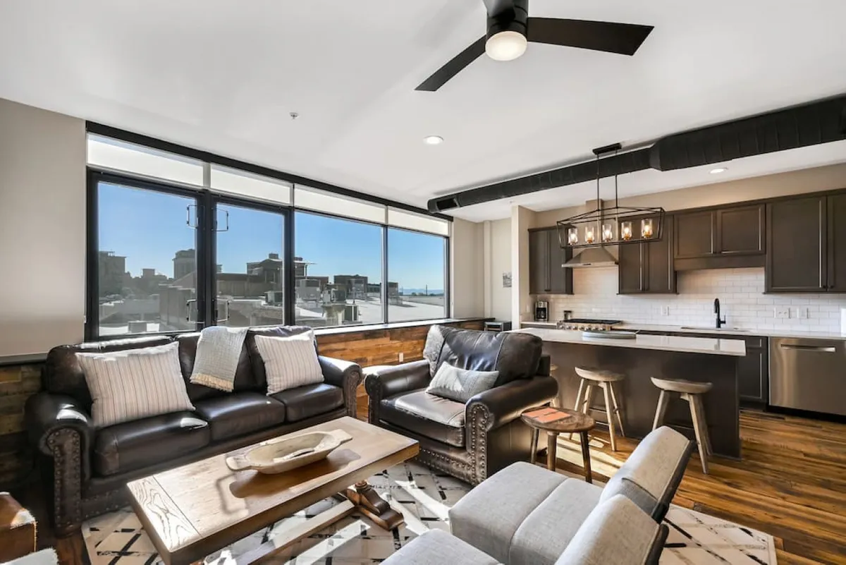 A stylish, open-plan living area with comfortable seating arranged around expansive windows that frame lively city views. The adjacent modern kitchen and urban skyline glimpsed outside reinforce the upscale, downtown ambiance.
