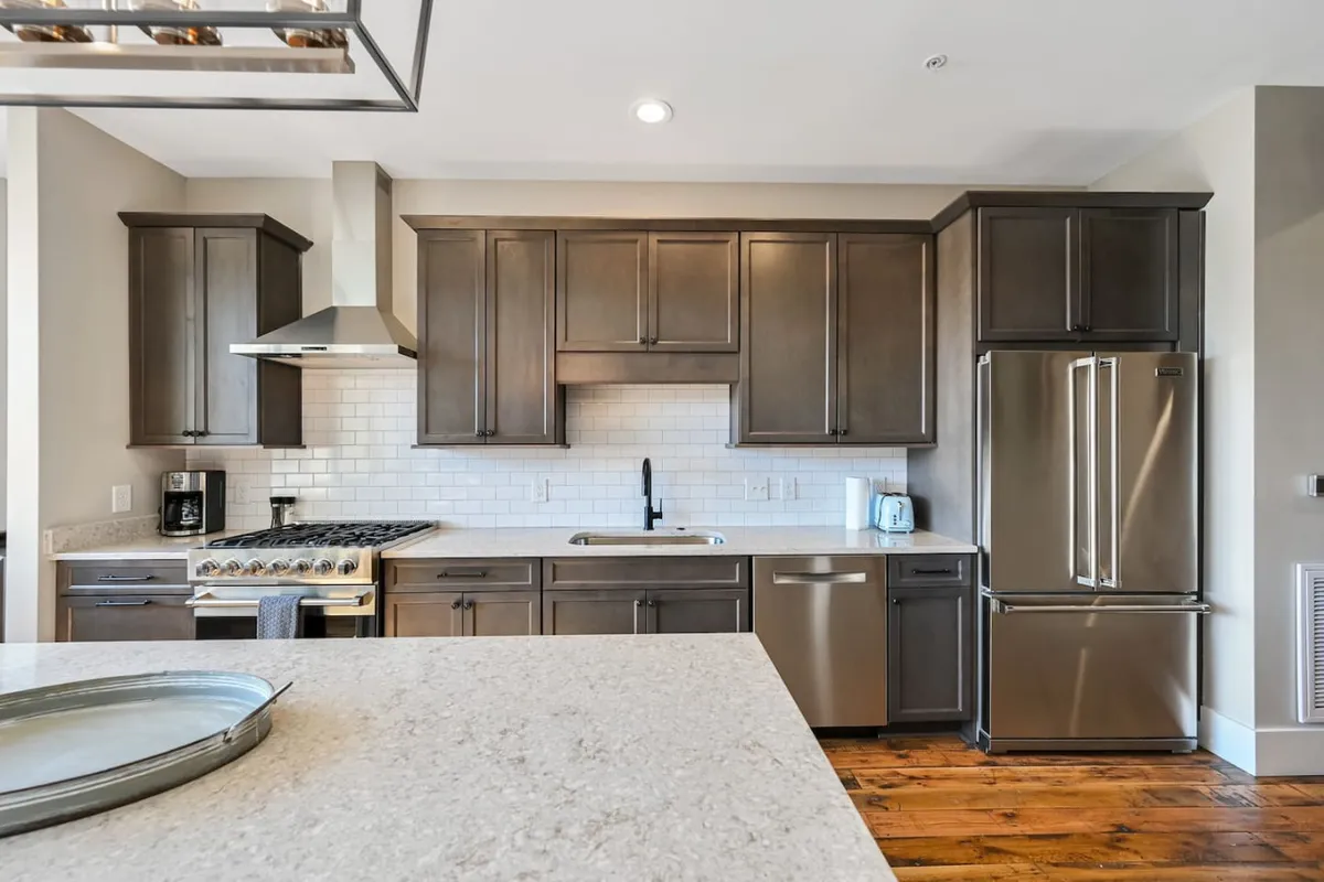 This image captures a warm, welcoming kitchen space featuring a central island, sleek stainless steel appliances, dark cabinetry, and a spacious countertop. The layout suggests a modern, comfortable environment well-suited for meal prep and casual gatherings.